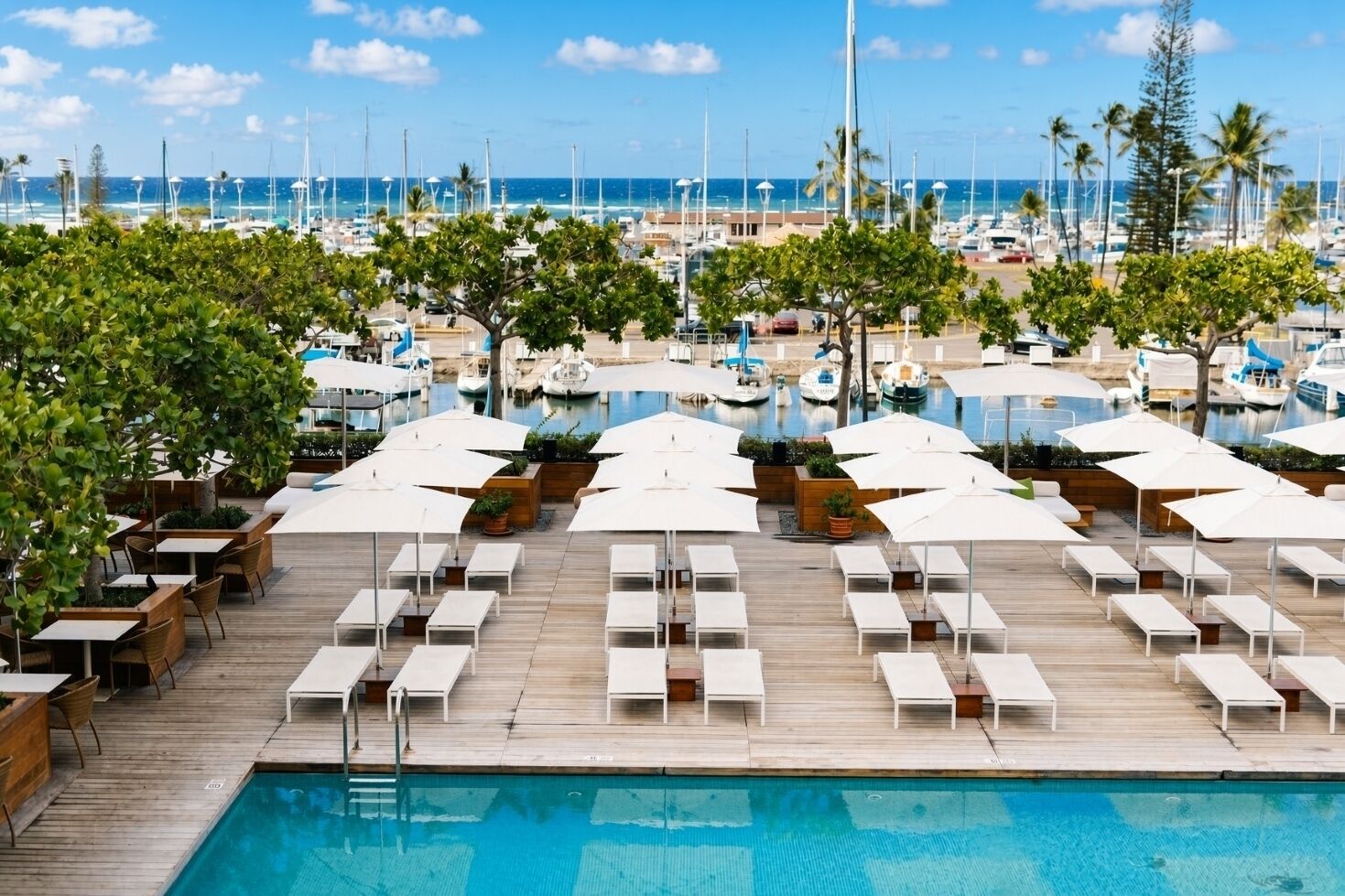 A poolside area with lounge chairs and umbrellas, a wooden deck, and a view of the marina with boats.