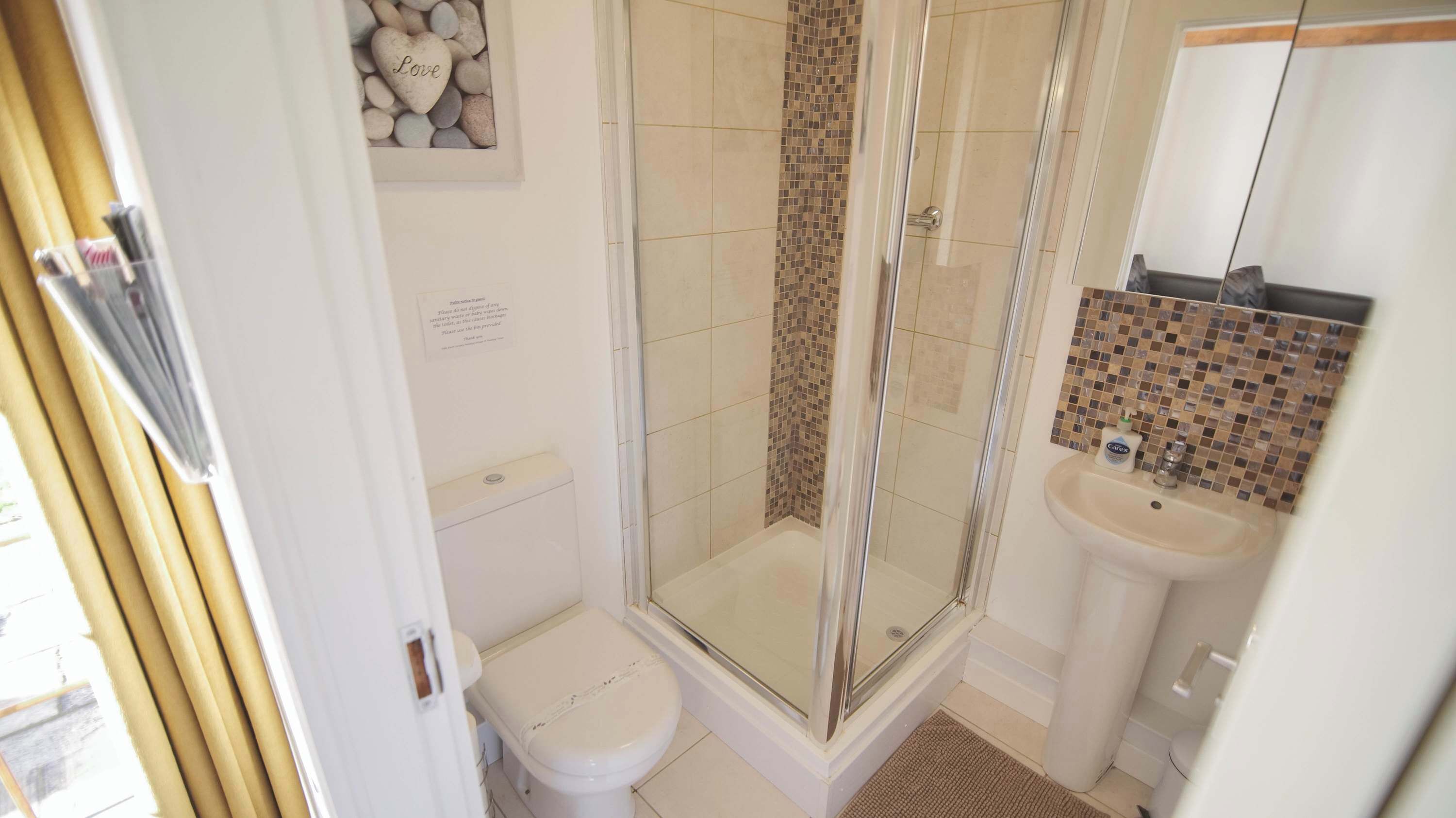 Et badeværelse med en bruseniche bag glaslåge, toilet og vask med mosaikfliser som baggrund.