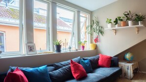 A modern living room with a dark blue sofa, red and blue cushions, a small side table with a globe, and a window with a view of a building outside. - Spacious, High-End Loft Apartment in the City Center (Unknown)