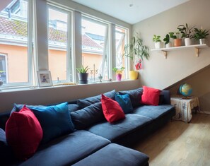 A modern living room with a dark blue sofa, red and blue cushions, a small side table with a globe, and a window with a view of a building outside. - Spacious, High-End Loft Apartment in the City Center (Unknown)