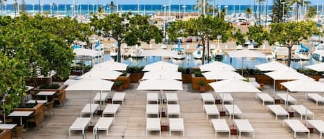 A poolside area with lounge chairs and umbrellas, a wooden deck, and a view of the marina with boats.