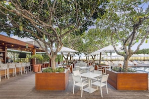 Un área de comedor al aire libre con mesas y sillas de madera, rodeada de árboles y sombrillas.