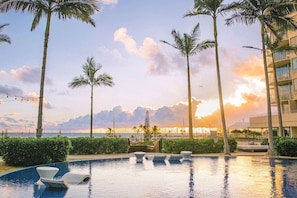 Zona de piscina con sillas de descanso, palmeras y vista al océano al atardecer.