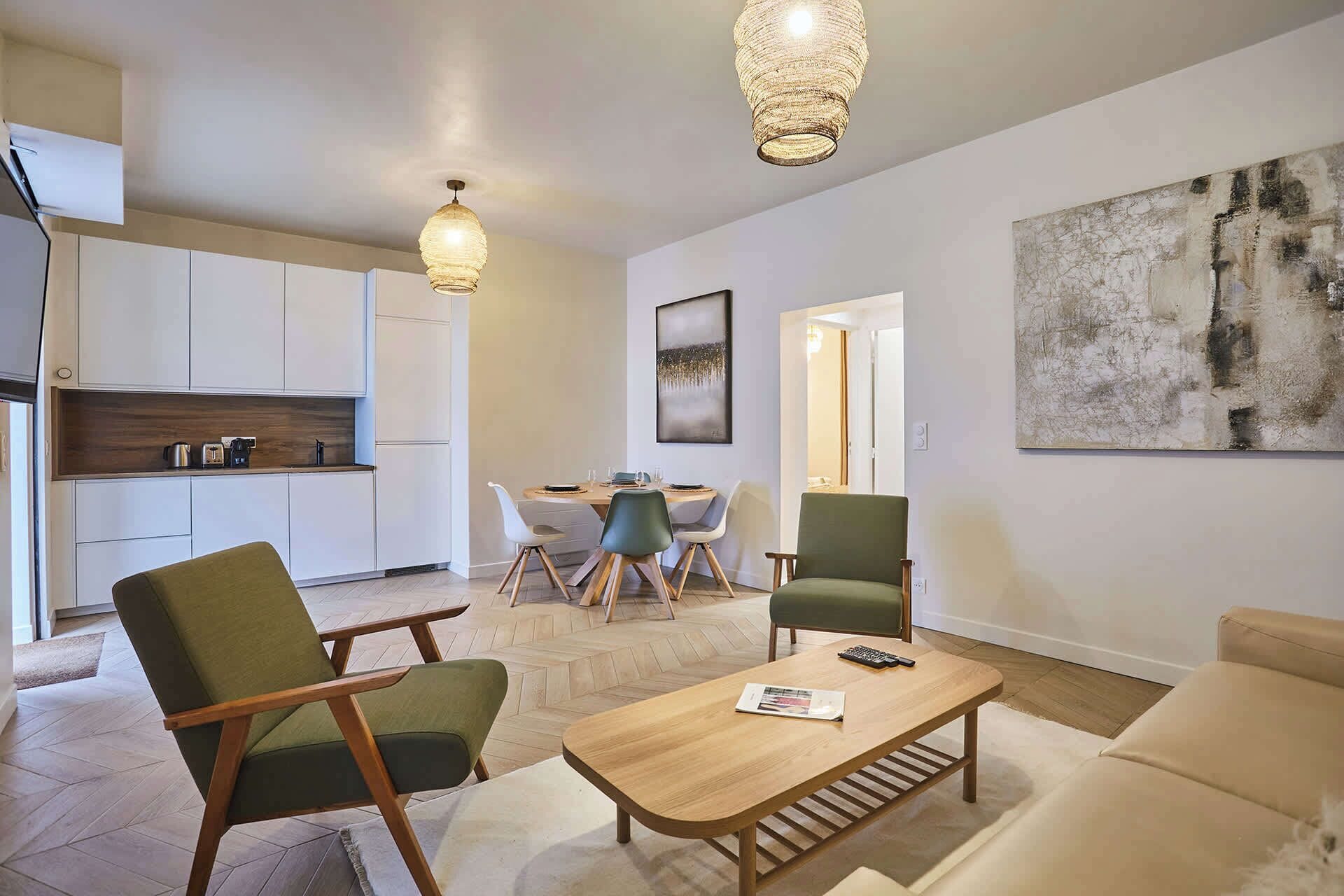 Un salon moderne avec une table basse en bois, des chaises vertes et blanches, et un canapé beige. On y trouve également un coin cuisine avec des meubles blancs et un plan de travail en bois. Deux lampes suspendues sont fixées au plafond.