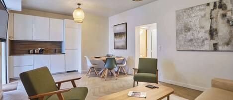 Un salon moderne avec une table basse en bois, des chaises vertes et blanches, et un canapé beige. On y trouve également un coin cuisine avec des meubles blancs et un plan de travail en bois. Deux lampes suspendues sont fixées au plafond.