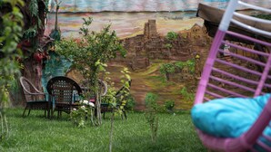 A rustic outdoor seating area with wicker chairs and a table, surrounded by greenery and a mural.