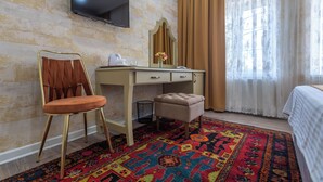 A hotel room with a bed, a desk with a chair, a television mounted on the wall, a mirror, a small trash bin, and a colorful rug.