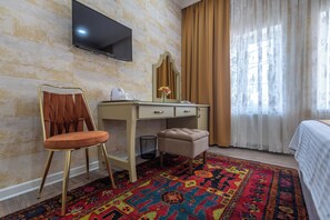 A hotel room with a bed, a desk with a chair, a television mounted on the wall, a mirror, a small trash bin, and a colorful rug.