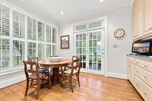 Un coin repas avec une table ronde et des chaises en bois, une grande fenêtre avec des stores, une porte à panneaux de verre et des meubles de rangement en bois.
