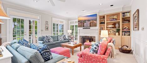 A spacious living room with a fireplace, a ceiling fan, and a large window overlooking a garden.