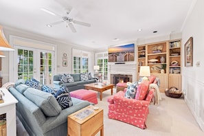 A spacious living room with a fireplace, a ceiling fan, and a large window overlooking a garden. - Fore Seasons Wonderful 5BR home in DeBordieu w/ super location, great porches (Georgetown)