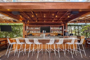 An outdoor bar area with a wooden deck, white chairs, and a bar stocked with bottles.