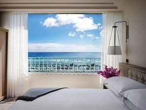 Een hotelkamer met een bed, nachtkastje, lamp, raam met uitzicht op zee en balkon met een hekwerk.