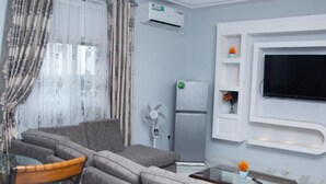 A living room with a grey sofa set, a small table with a vase, a flat-screen TV mounted on the wall, and a white air conditioning unit. - Champion Hotel (DOUALA)