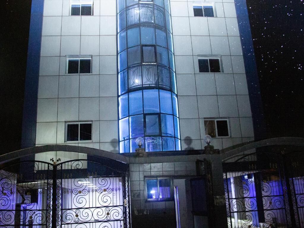 Un immeuble à plusieurs étages doté d’un ascenseur à cage de verre central, d’une entrée fermée par un portail orné de ferronnerie décorative et offrant une vue sur un ciel nocturne dégagé.