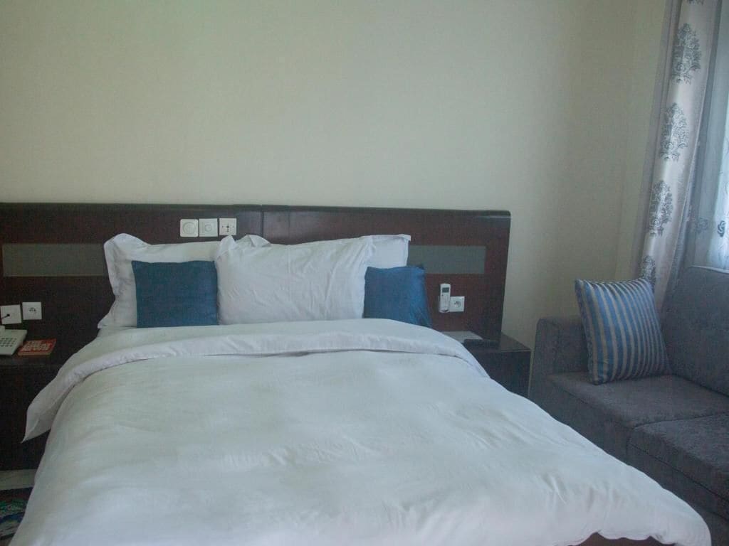 Quarto de hotel com cama, mesa de cabeceira, luminária, sofá e janela com cortinas.