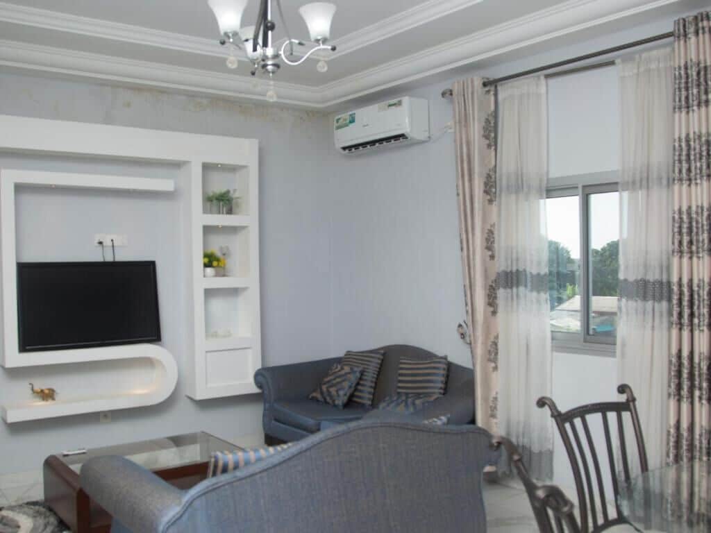 Una sala de estar con un televisor de pantalla plana montado en una pared blanca, un sofá gris con cojines estampados, una mesa de centro de vidrio y una silla de madera.