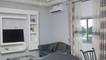 Una sala de estar con un televisor de pantalla plana montado en una pared blanca, un sofá gris con cojines estampados, una mesa de centro de vidrio y una silla de madera.