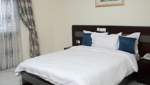 A neatly made bed with white bedding, a dark headboard, and a bedside table with a telephone. - Champion Hotel (DOUALA)
