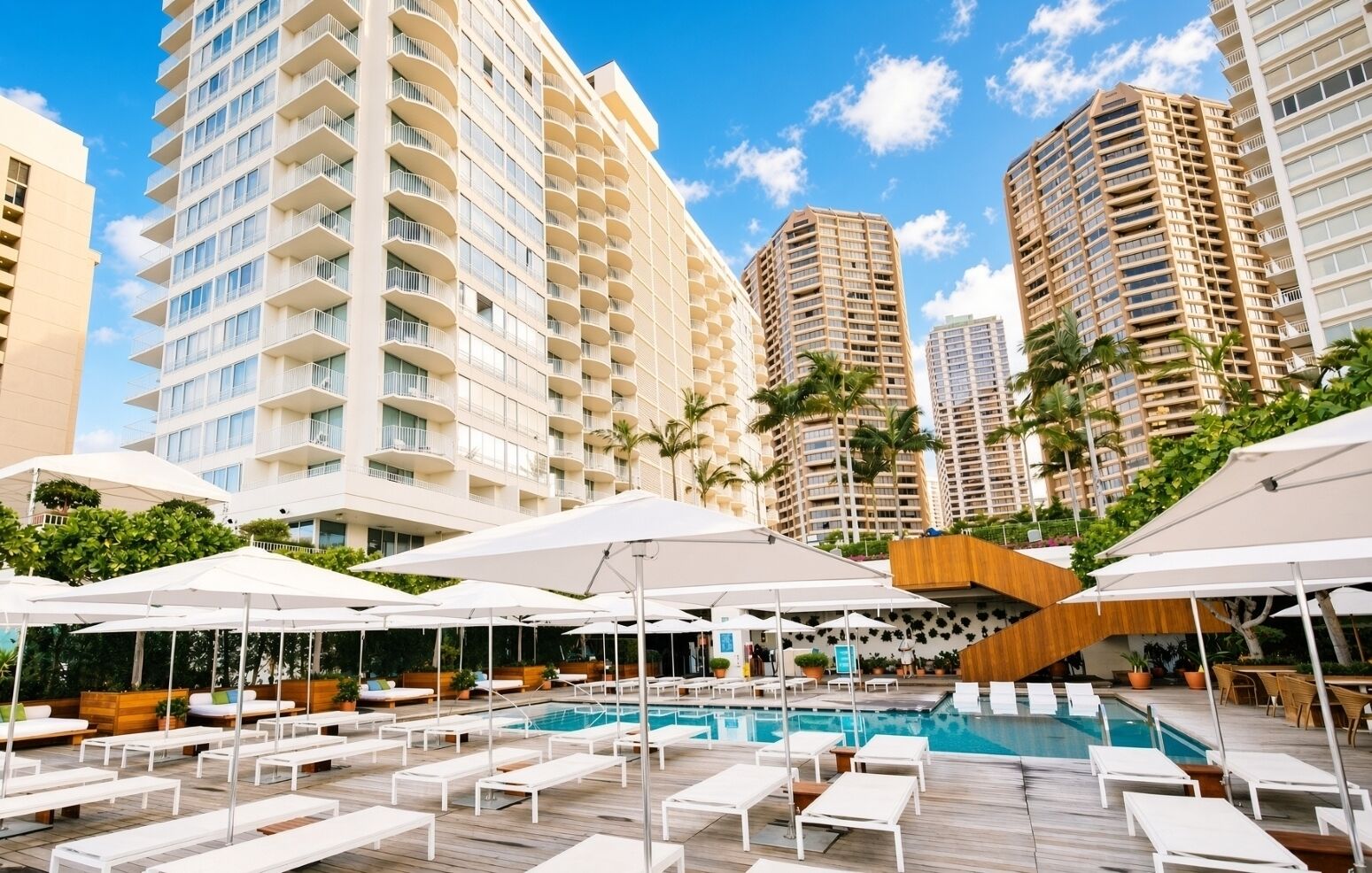 Zona de piscina hotelera con tumbonas y sombrillas, rodeada de edificios altos.