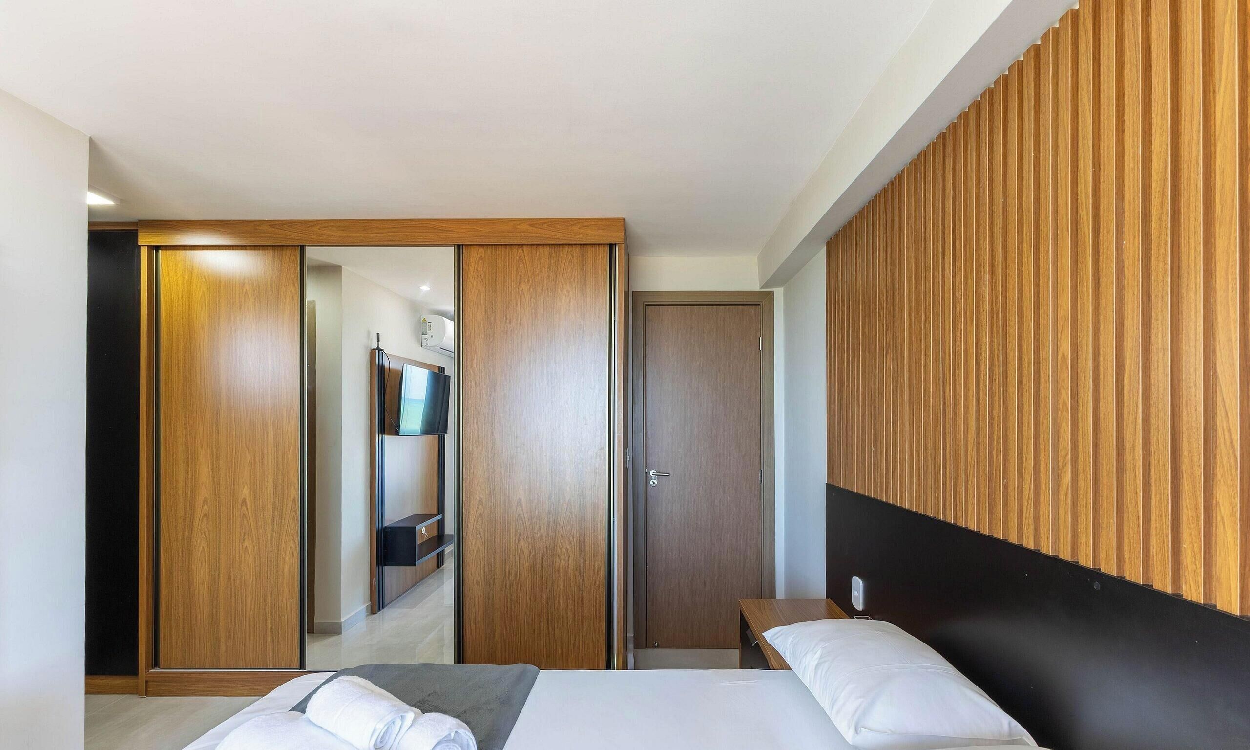 A modern hotel room with a bed, a wooden panel wall, a wardrobe, and a bathroom with a mirror and towel rack.