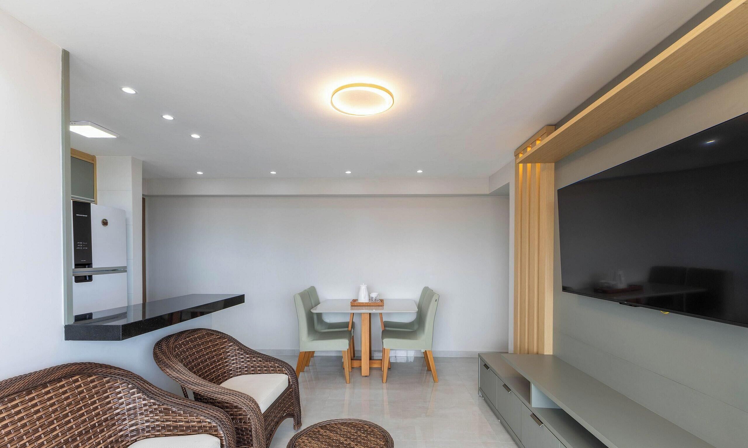 A modern living room with a flat-screen TV, wicker furniture, a dining table, and recessed ceiling lighting.