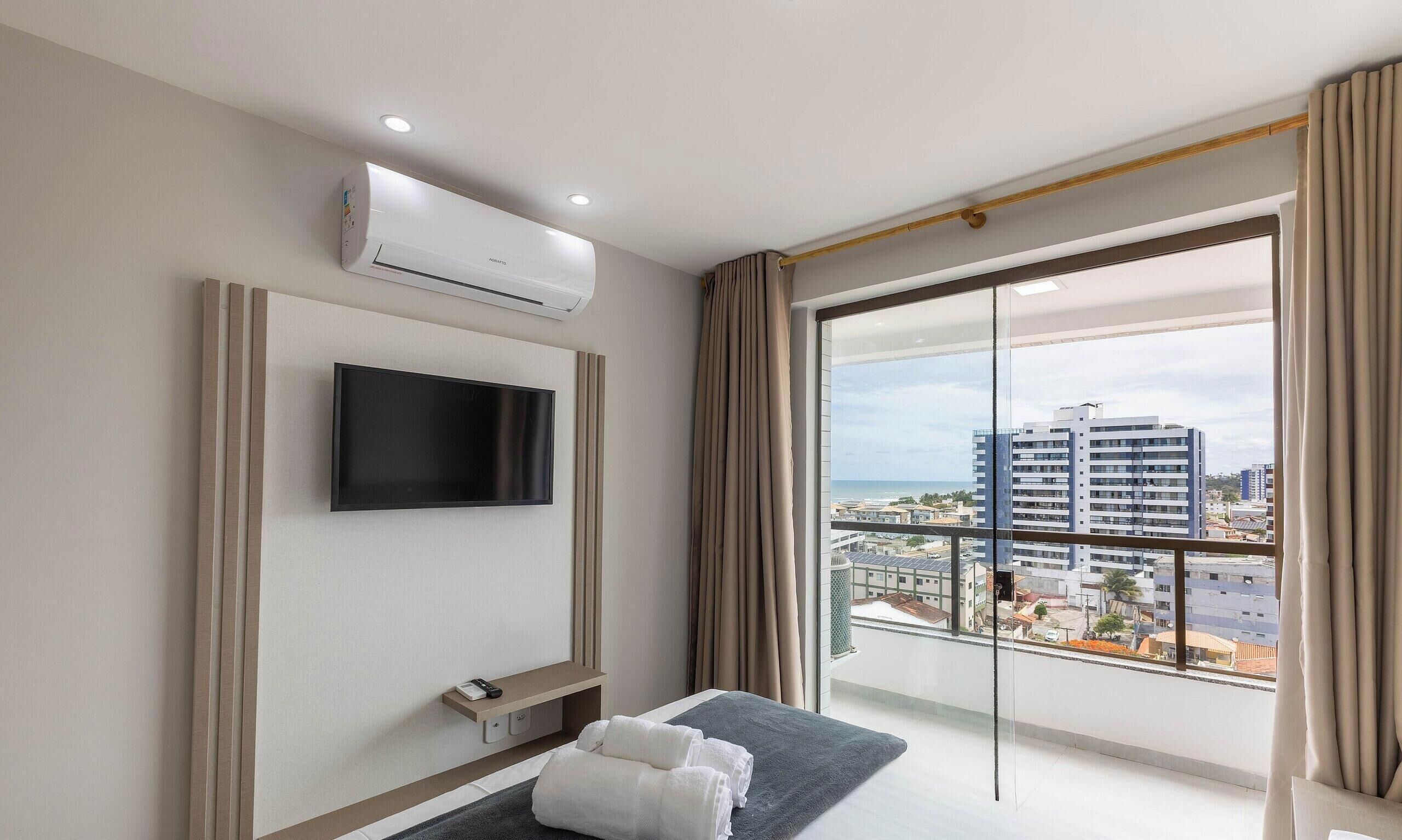 A hotel room with a flat-screen TV mounted on the wall, a small bedside table with a remote control, a bench with towels, and a large window offering a city view.