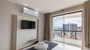 A hotel room with a flat-screen TV mounted on the wall, a small bedside table with a remote control, a bench with towels, and a large window offering a city view. - Apt Sea View 300 m From the Beach Mmd0802 (Ilhéus)