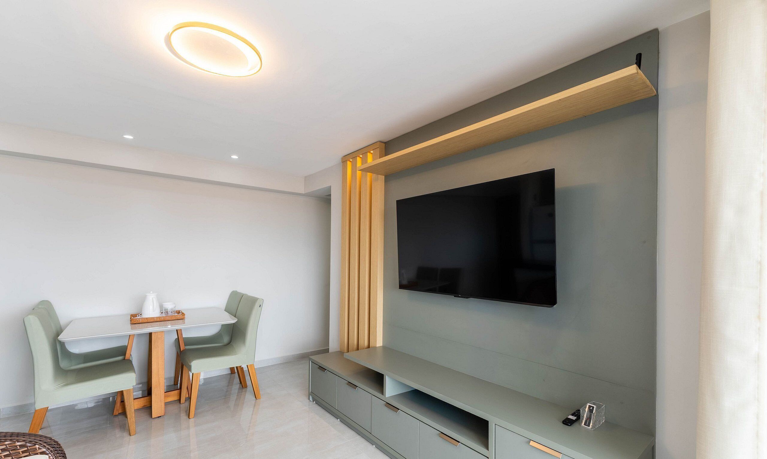 A modern living room with a flat-screen TV mounted on a grey wall, a wooden shelf below, and a dining area with a table and chairs.
