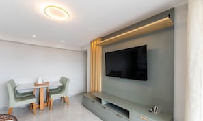 Una sala moderna con un televisor de pantalla plana montado en una pared gris, un estante de madera debajo y una zona de comedor con una mesa y sillas.