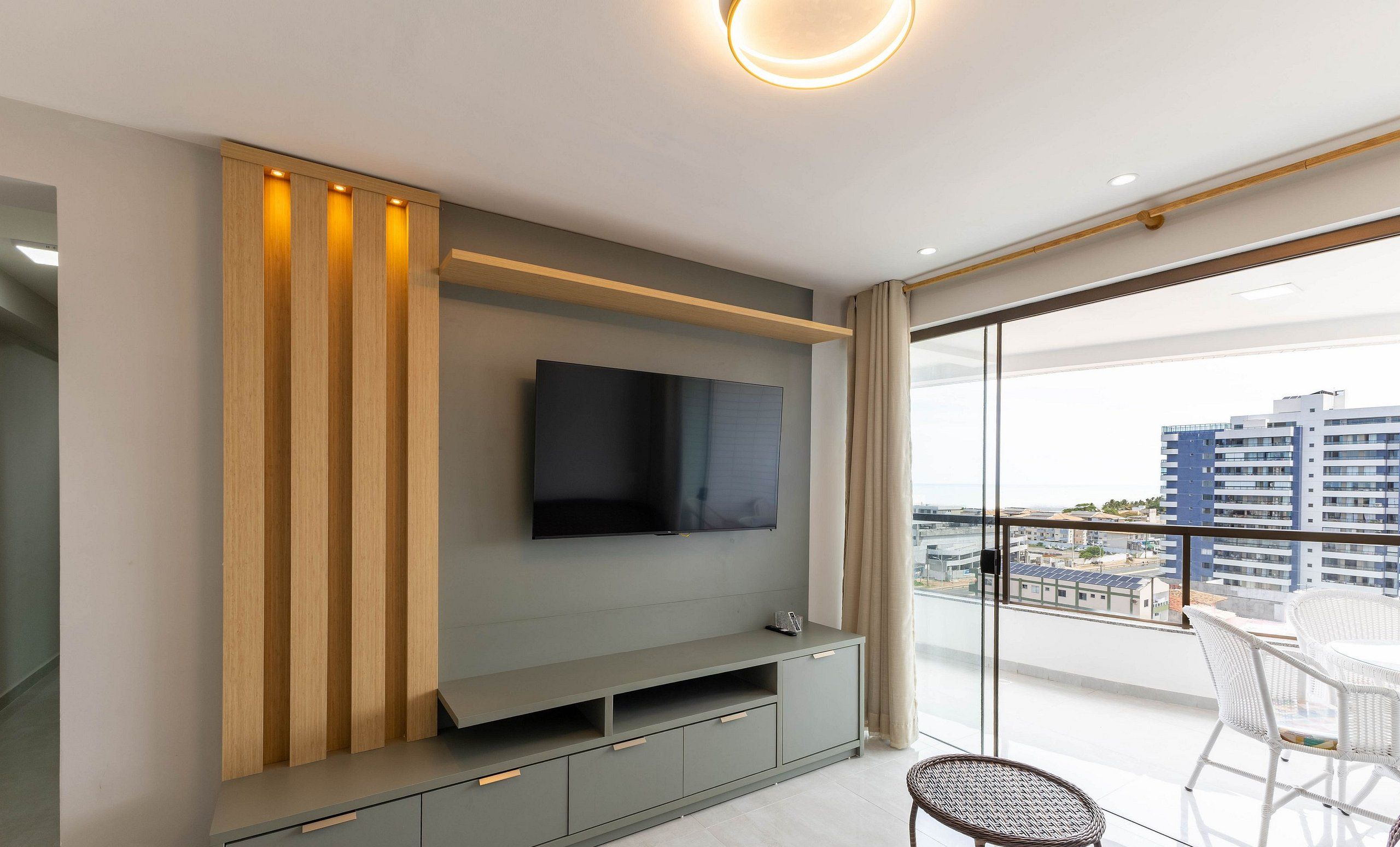 A modern living room with a flat-screen TV mounted on a grey wall, wooden slats on the left, and a balcony with white wicker chairs.