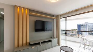 A modern living room with a flat-screen TV mounted on a grey wall, wooden slats on the left, and a balcony with white wicker chairs.