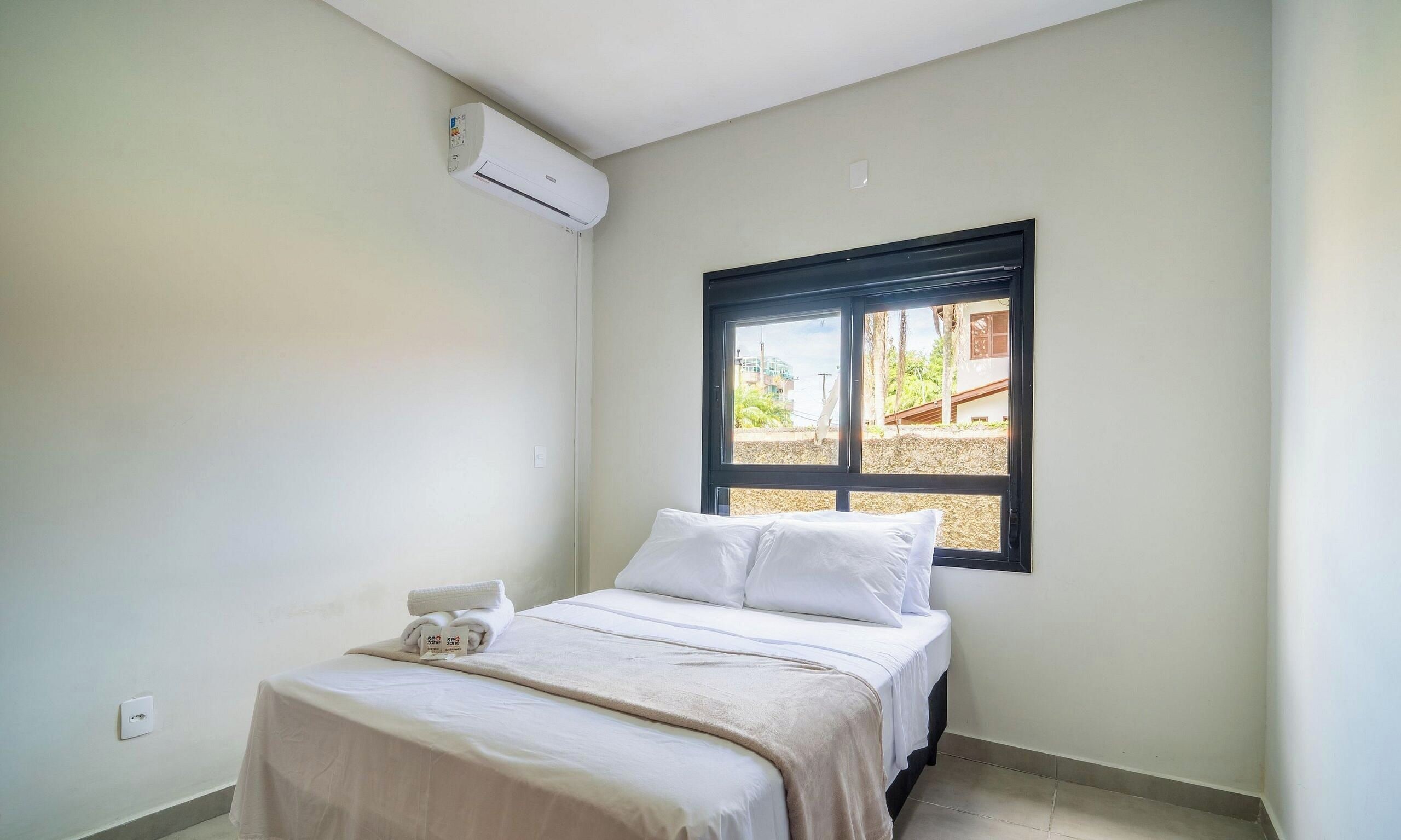 A small, single-bed room with a window, an air conditioning unit, and a neatly made bed.