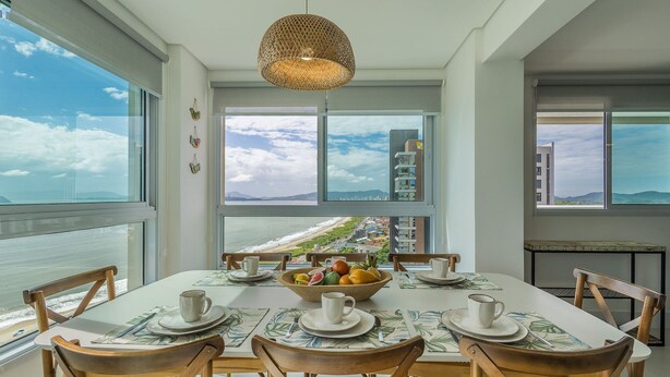 Comedor con una mesa preparada para cuatro, un frutero y vista al océano y edificios.