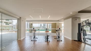 Un gimnasio con equipamiento de ejercicio, ventanas amplias y vistas a edificios.