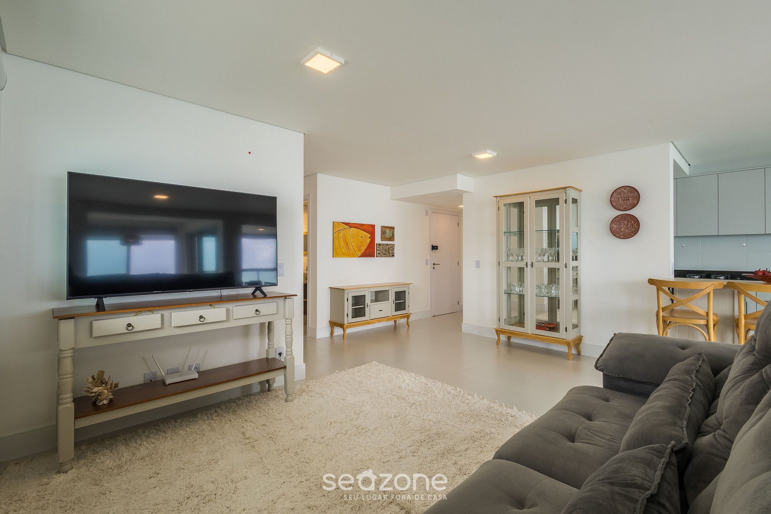 Una sala de estar moderna con un televisor de pantalla plana, un sofá gris, una consola blanca y un aparador de vidrio.