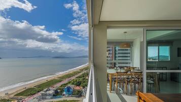 A balcony with a view of the beach and ocean, a dining area with chairs and a table, and a hanging light fixture.