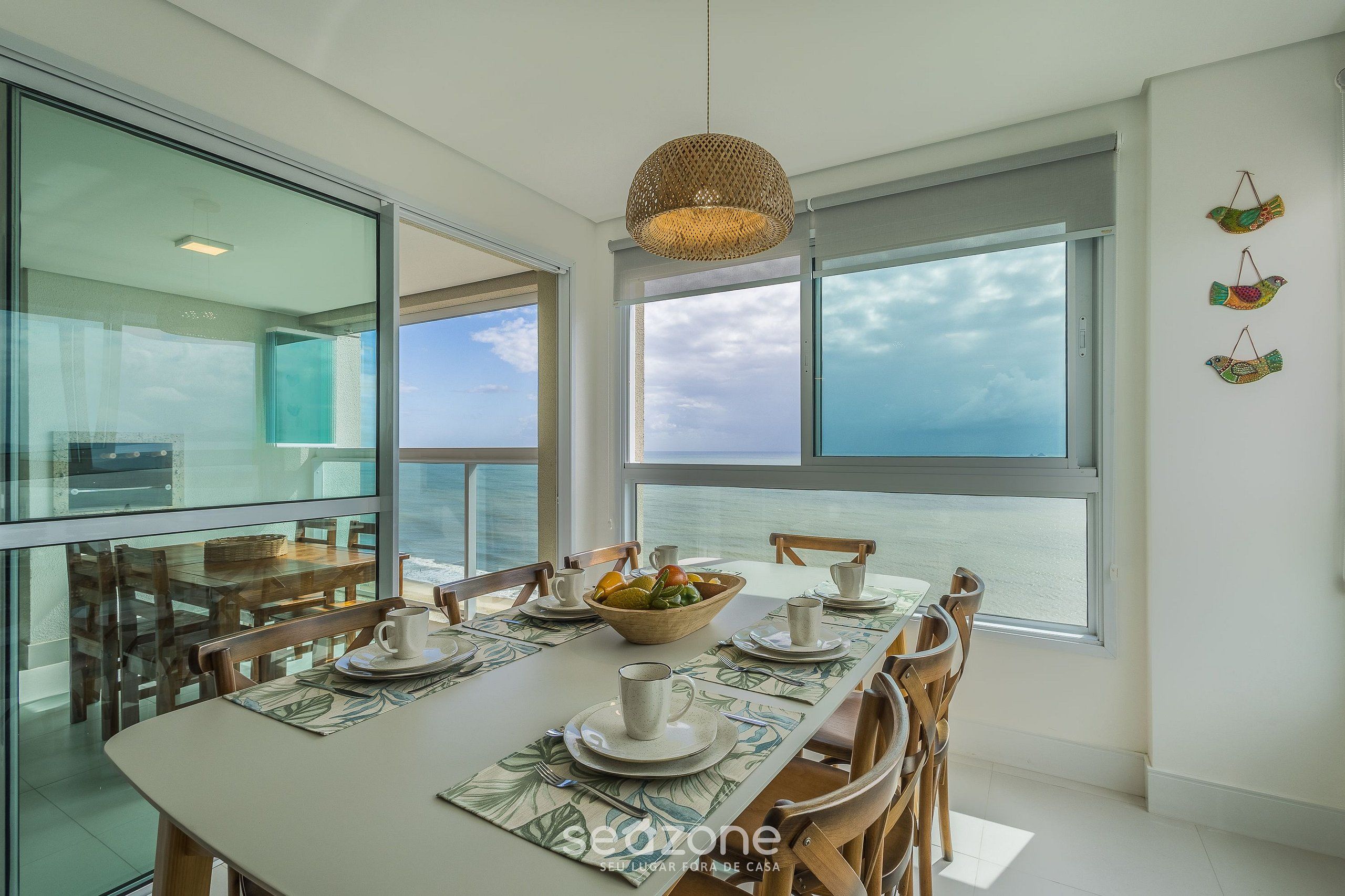 Uma sala de jantar com uma mesa grande posta para quatro pessoas, uma luminária pendente trançada e vista para o mar através de grandes janelas.