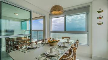 Uma sala de jantar com uma mesa grande posta para quatro pessoas, uma luminária pendente trançada e vista para o mar através de grandes janelas.
