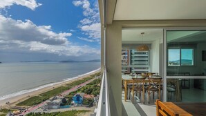 Un balcón con vista a la playa y al océano, un área de comedor con sillas y una mesa, y un lámpara colgante.