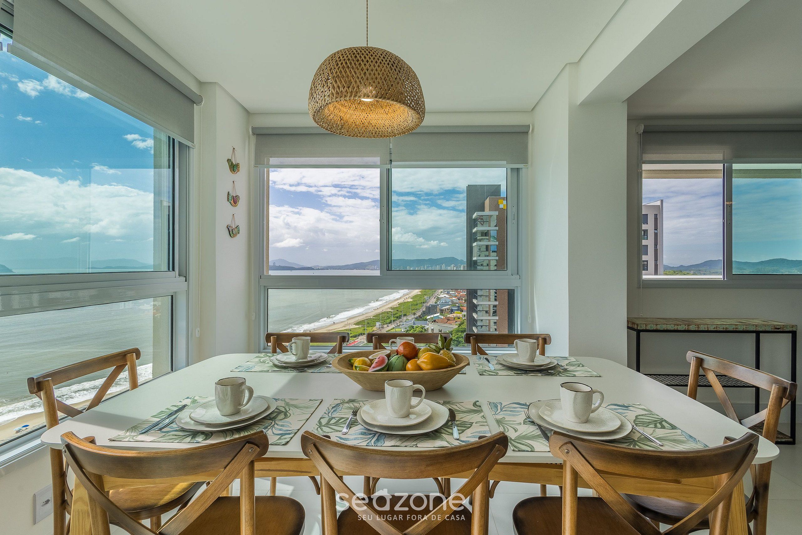 Uma sala de jantar com uma mesa posta para quatro pessoas, uma tigela de frutas e vista para o oceano e edifícios.