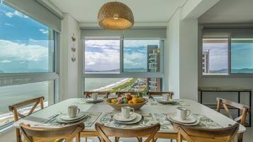 Uma sala de jantar com uma mesa posta para quatro pessoas, uma tigela de frutas e vista para o oceano e edifícios.