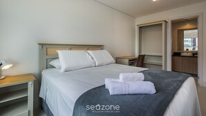 Una cama bien hecha con sábanas blancas y una manta gris, una mesita de noche con una lámpara y una mesita auxiliar de madera.