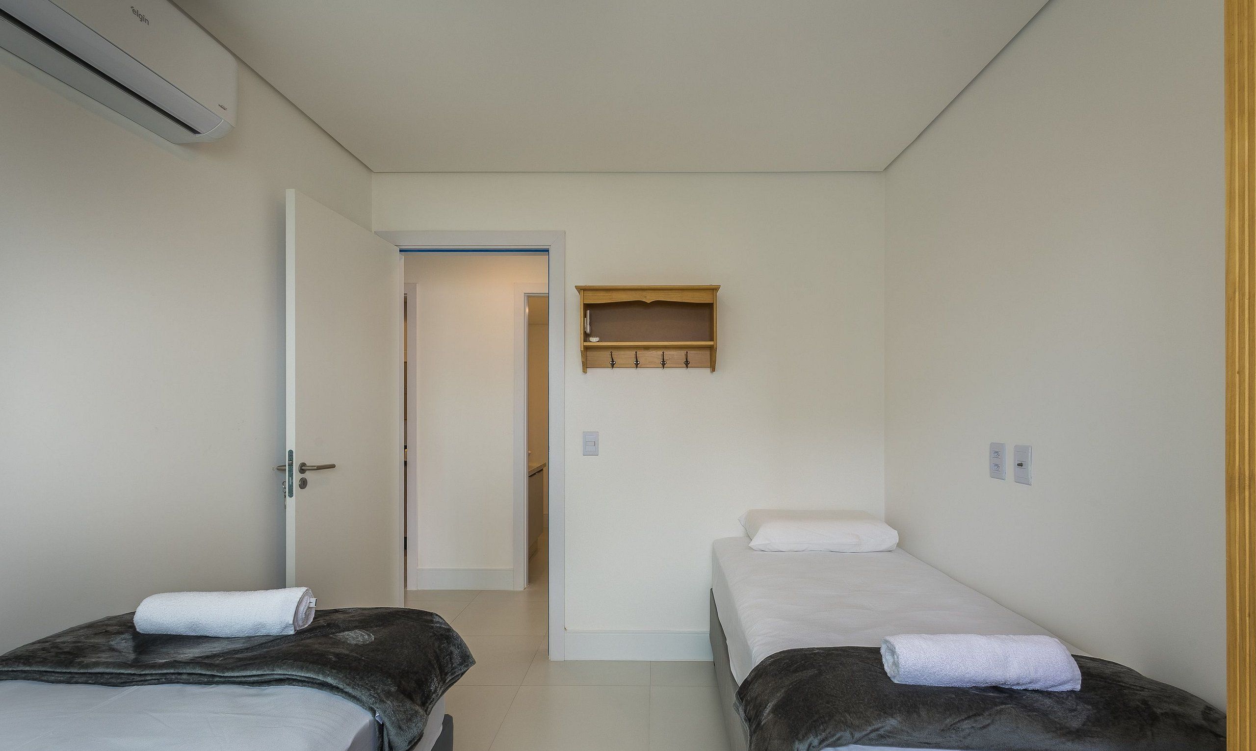 Una habitación pequeña y limpia con dos camas, un estante de madera sobre la puerta y una pared blanca.