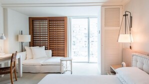 Quarto de hotel com cama, escrivaninha com luminária, cadeira, mesa pequena e uma janela grande com persianas.