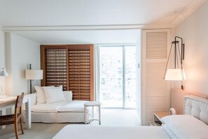 Habitació d'hotel amb llit, escriptori amb làmpada, cadira, taula petita i finestra gran amb persianes.