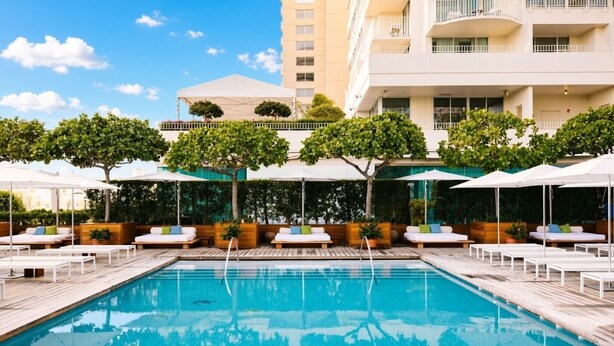Piscina com espreguiçadeiras e guarda-sóis, cercada por vegetação e um edifício com vários andares ao fundo.