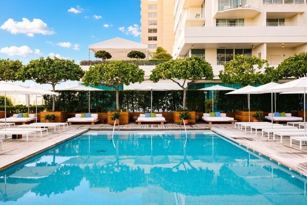 Una piscina amb gandules i ombrelles, envoltada de vegetació i un edifici de diversos pisos al fons.