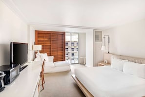 Une chambre d’hôtel équipée d’un lit, d’une télévision, d’un bureau avec un ordinateur, d’une chaise et offrant une vue sur la ville grâce à une grande fenêtre.