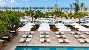 Área da piscina com espreguiçadeiras e guarda-sóis, um deck de madeira e vista para a marina com barcos.